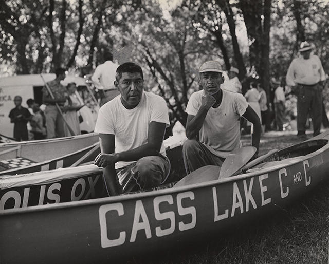 Two Ojibwe men crouch in a canoe with the words "Cass Lake" written on it in white capital letters.